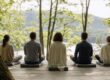 Five people sit in a row on cushions, meditating with their backs to the camera on a wooden deck overlooking a serene lake and forest.